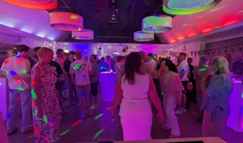 Mensen dansen en praten op een feest in een zaal met kleurrijke discoverlichting en hoge tafels.